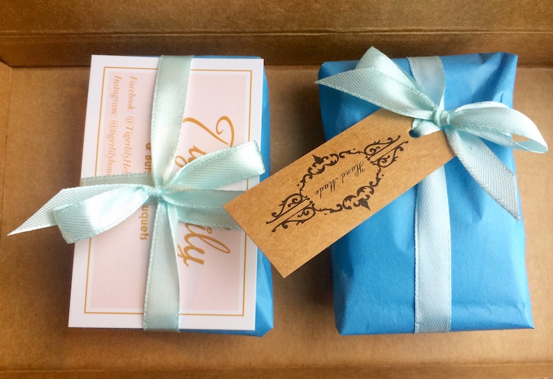 Puede incluir: Dos peque&ntilde;os regalos envueltos en papel de seda azul con cinta azul claro y una etiqueta marr&oacute;n que dice "Hecho a mano". Un regalo es una tarjeta con fondo rosa y el texto "Realmente un hermoso ramo" y el otro regalo es una peque&ntilde;a caja.