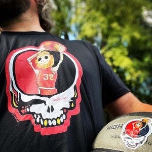May include: A black t-shirt with a red and yellow graphic of a skeleton playing basketball. The graphic is inside a red and yellow skull design. The text "Grateful Dead" is printed on the shirt. The shirt is being held by a person who is also holding a basketball.