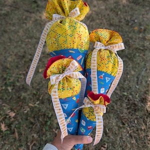 May include: Four cone-shaped fabric gift bags in varying sizes, featuring a yellow top section with a multi-colored dot pattern and a blue bottom section with school supply graphics. Each bag is tied with a white ribbon printed with pencil icons.