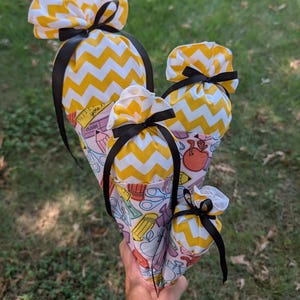 May include: Four cone-shaped fabric gift bags in varying sizes, featuring a yellow and white chevron pattern at the top and a school supply print on the bottom. Each bag is tied with a black satin ribbon. The school supply print includes pencils, scissors, apples, and rulers with the text "you erase".