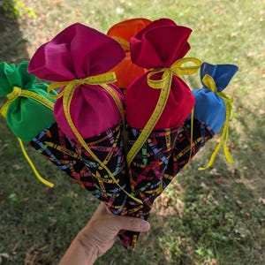 May include: A collection of five cone-shaped fabric gift bags featuring a black base with a colorful crayon print. Each bag is topped with a solid-colored fabric in green, pink, orange, red, or blue, tied with a yellow measuring tape ribbon.