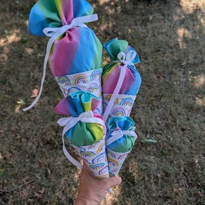 May include: Four cone-shaped fabric gift bags in varying sizes, featuring a white base with a colorful rainbow pattern and a top section of tie-dye fabric in blue, pink, and green. Each bag is cinched at the center with a white ribbon bow.