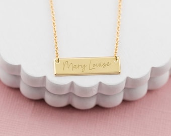 May include: A gold-toned metal bar necklace featuring a delicate chain and a rectangular pendant engraved with the name Mary Louise in a cursive script font. The piece is suspended by two thin gold-toned links attached to the top corners of the bar.