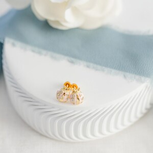 May include: A pair of gold-colored earrings with a pink enamel finish and clear crystal accents. The earrings are shaped like small ornaments and are displayed on a white, textured surface with a blue ribbon and a white flower in the background.