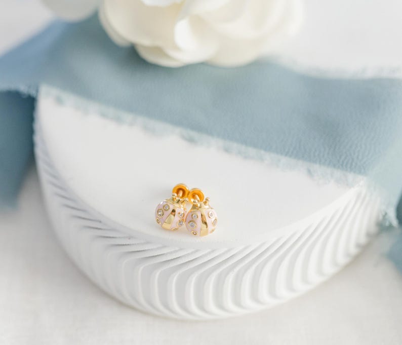 May include: A pair of gold-colored earrings with a pink enamel finish and clear crystal accents. The earrings are shaped like small ornaments and are displayed on a white, textured surface with a blue ribbon and a white flower in the background.