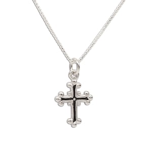 Peut inclure: Un pendentif en forme de croix en métal argenté suspendu à une chaîne maille carrée assortie. La croix présente un design ornemental avec des extrémités arrondies et bombées aux quatre points et un détail central sombre et creusé.