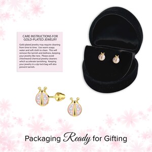 May include: Gold-plated ladybug stud earrings with pink enamel and clear crystal accents. The earrings are displayed in a black velvet jewelry box. The image also includes care instructions for gold-plated jewelry and the text "Packaging Ready for Gifting."