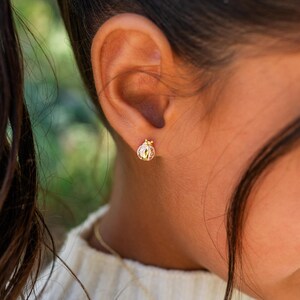May include: A close-up of a gold ladybug stud earring. The earring is detailed with a red and white design on the body and a gold crown on the head. The earring is worn on a person's ear.