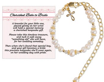 May include: A gold-toned bracelet with white pearls and a small cross charm. The bracelet is displayed with a card that reads "Cherished Babe to Bride" and includes text about a keepsake for a bride.