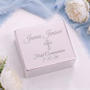 May include: A silver-colored, square box with the engraved text "James Jenson", a cross, and "First Communion 7-12-26". The box is surrounded by white flowers and a light blue fabric.