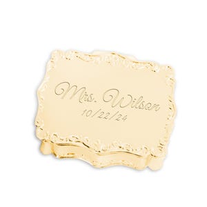 May include: A rectangular gold-toned metal trinket box with an ornate, scalloped edge. The lid is engraved with the text "Mrs. Wilson" in a cursive script font, followed by the date "10/22/24" in a simple, clean, sans-serif typeface.
