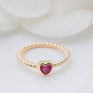 May include: A gold-colored ring with a twisted band design. The ring features a heart-shaped, ruby-red gemstone at the center. The ring is set against a soft, white background.