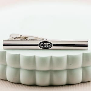 Könnte beinhalten: Silberne Krawattenklammer mit einem schwarzen Streifen und einem schwarzen Oval mit den Initialen CTR in Weiß.