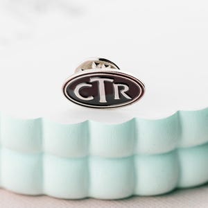 Könnte beinhalten: Eine ovale silberne Anstecknadel mit schwarzem Emaille-Hintergrund und den Buchstaben "CTR" in Weiß.