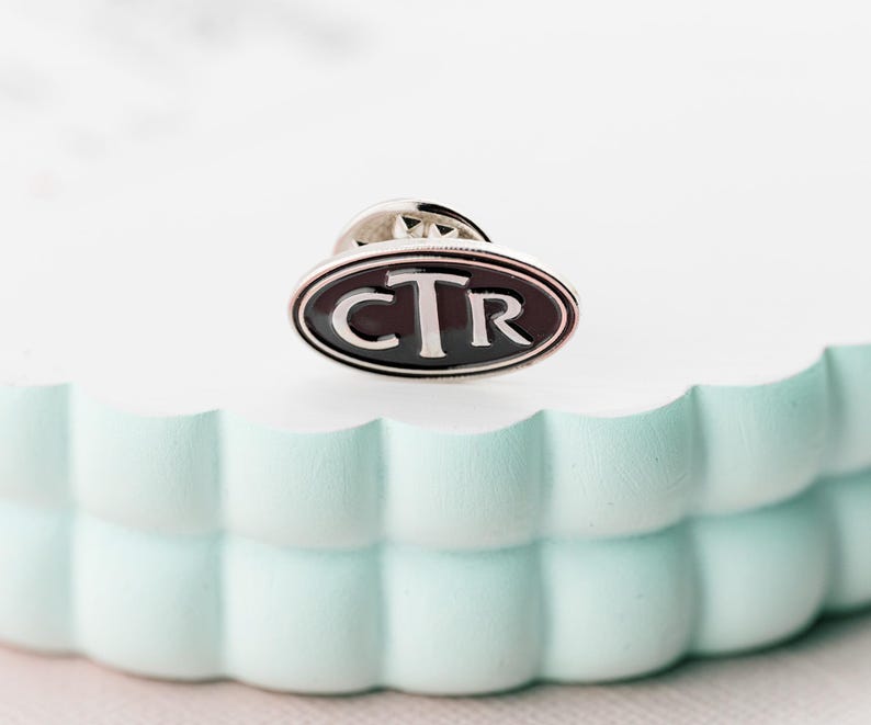Puede incluir: Un pin ovalado de plata con fondo de esmalte negro y las letras "CTR" en blanco.