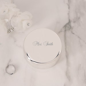 May include: A round, polished silver-tone metal trinket box featuring the engraved text "Mrs. Smith" in a cursive script on the lid. The container is accompanied by a simple silver-tone ring and a white floral accessory with small clear crystals.