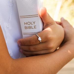 May include: A white "Holy Bible" book is held in the hands. A silver ring with a heart-shaped design and small stones is on the finger. The ring has the initials "CTR" engraved on it.