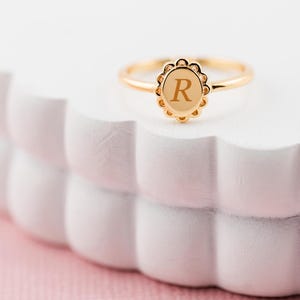 May include: A gold ring with an oval face displaying the letter "R". The ring has a scalloped edge. It is set against a white, textured background. The ring is a piece of jewellery.