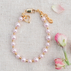 Peut inclure: Un bracelet composé de perles rose clair alternant avec de petites perles dorées. Le bijou comprend un fermoir mousqueton doré, une breloque en forme de cœur avec des pierres transparentes et une étiquette ronde gravée de la lettre S.