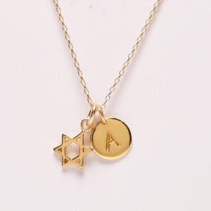May include: A gold-toned necklace featuring a delicate chain with two charms attached. One charm is a six-pointed Star of David, and the other is a small, circular disc engraved with the capital letter A in a clean, sans-serif font style.