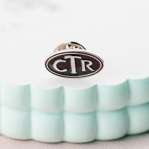 Könnte beinhalten: Eine ovale silberne Anstecknadel mit schwarzem Emaille-Hintergrund und den Buchstaben "CTR" in Weiß.