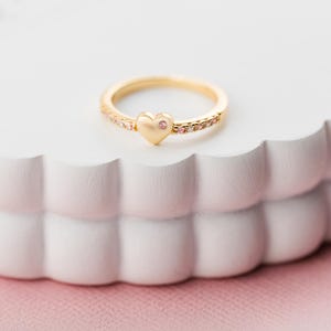 May include: A gold-colored ring with a heart-shaped center stone and small pink gemstones. The ring is set on a white, scalloped surface, with a pink background. The ring is a piece of jewelry.