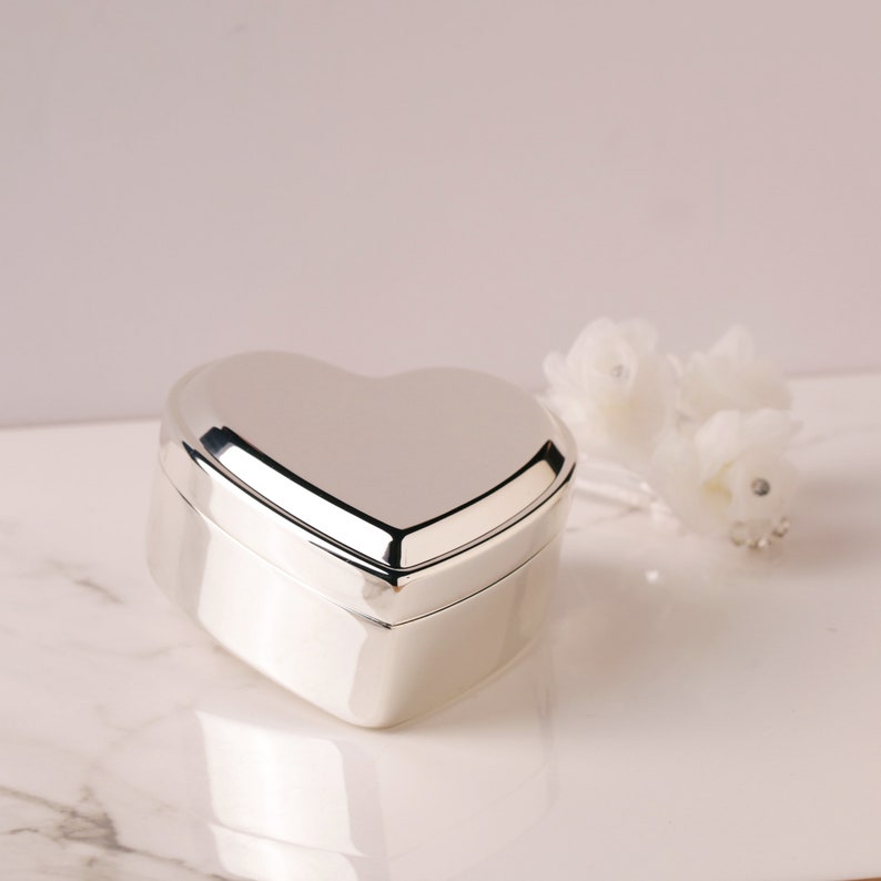 May include: A silver heart-shaped trinket box with a smooth, polished finish. The box is closed and sitting on a white marble surface.