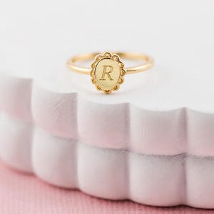 May include: A gold ring featuring an oval-shaped face with a scalloped edge. The center of the oval is engraved with the capital letter R. The band is thin and polished, creating a simple and elegant design for this personalized jewelry piece.