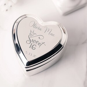 May include: Silver heart-shaped trinket box with an engraved design that says "Olivia Mae Sweet 16 5.13.20".