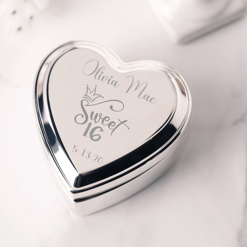 May include: Silver heart-shaped trinket box with an engraved design that says "Olivia Mae Sweet 16 5.13.20".