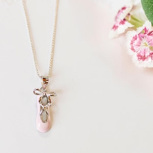 Peut inclure: Un collier en argent avec un pendentif en forme de ballerine rose. La chaussure a un nœud argenté et un contour argenté.