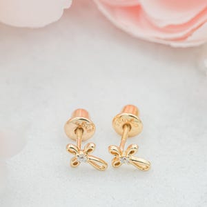 May include: A pair of gold-colored cross stud earrings with a small, clear gemstone at the center of each cross. The earrings have a screw-back closure. The background features soft pink and white tones.