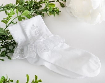 May include: A pair of white socks with a delicate lace trim and an embroidered cross detail. The socks are made of a soft, ribbed material and are designed for a special occasion. The socks are displayed on a white surface with greenery.