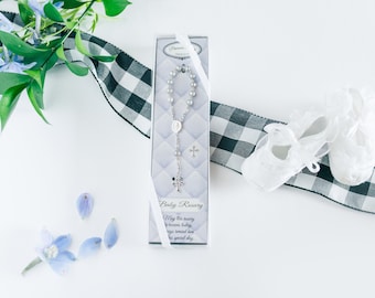 May include: A white box with a silver baby rosary inside. The box has a light blue and white design with the words "Baby Rosary" and a message about the rosary being a special gift. The box is sitting on a white surface with a black and white checkered ribbon and white baby shoes.