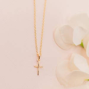 May include: A delicate gold-toned necklace featuring a small, simple cross pendant suspended from a thin, polished cable chain. The metal has a bright, reflective finish, and the cross design is minimalist with clean, straight edges and a smooth surface.