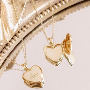 May include: Two gold heart-shaped locket necklaces on thin chains. One locket is closed and engraved with the name Ava in script font, while the other locket is open, revealing a hollow interior. Both pieces feature a polished metallic finish.