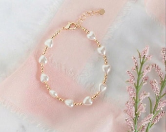 May include: A gold-toned bracelet featuring alternating small round metallic beads and white heart-shaped beads. The bracelet includes a lobster claw clasp and an adjustable chain extension with a small circular charm at the end.
