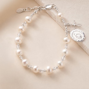 May include: A silver bracelet featuring white freshwater pearls and small clear crystals. The bracelet includes a silver lobster clasp, an adjustable chain with a small tag, and two dangling charms: a silver cross and an oval medal with an embossed figure.