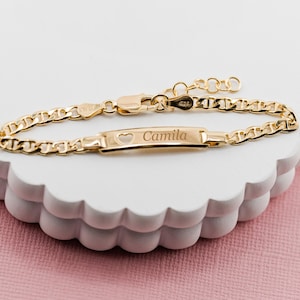 May include: A gold bracelet with a chain link design. The bracelet has a rectangular plate engraved with the name "Camila" and a heart-shaped cutout. It is displayed on a white, scalloped surface against a pink background.