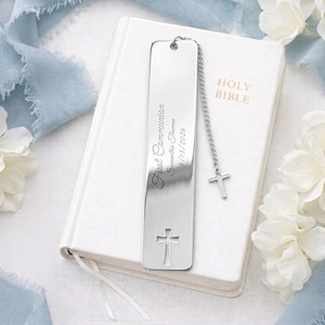 Op de afbeelding: Een zilveren boekenlegger met een kruisontwerp en de woorden "First Communion" gegraveerd. De boekenlegger is bevestigd aan een ketting met een kleine kruisbedel. Het rust op een wit boek met de woorden "Holy Bible" in gouden letters.