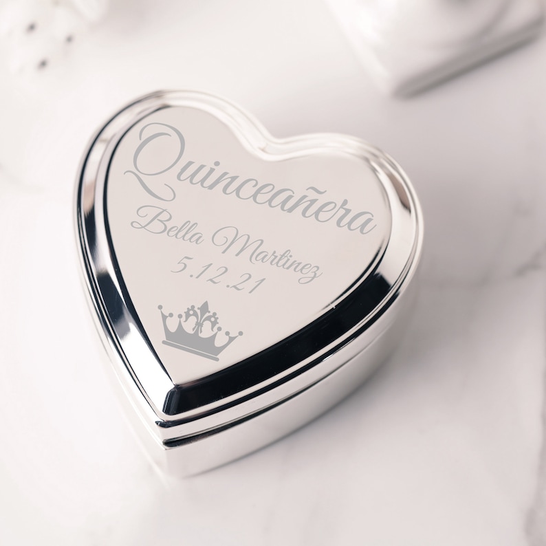 May include: Silver heart-shaped trinket box with an engraved inscription that reads "Quincea&ntilde;era Bella Martinez 5.12.21" and a crown design.