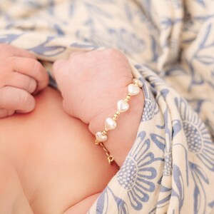 May include: A delicate bracelet featuring a series of small, white heart-shaped beads alternating with tiny, polished gold-toned metallic spheres. The bracelet includes a gold-toned chain link extension and a clasp, worn around a small wrist.