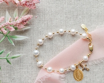 May include: A gold-toned bracelet featuring white pearls and small clear beads, accented with a gold-toned oval pendant depicting a religious figure and a small gold-toned cross charm with a single clear stone, finished with a lobster clasp.