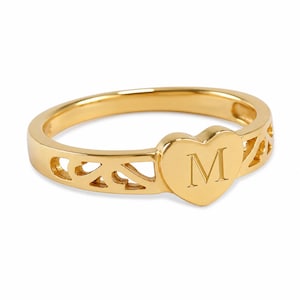 May include: A gold-toned ring featuring a central heart-shaped plaque engraved with the capital letter M. The band displays a decorative openwork cutout pattern along the sides, creating a continuous metal design with a polished finish.
