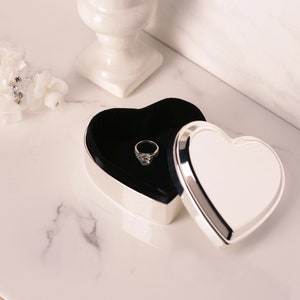 May include: A silver heart-shaped ring box with a black velvet interior. The box is open and a silver ring with a clear stone is inside.