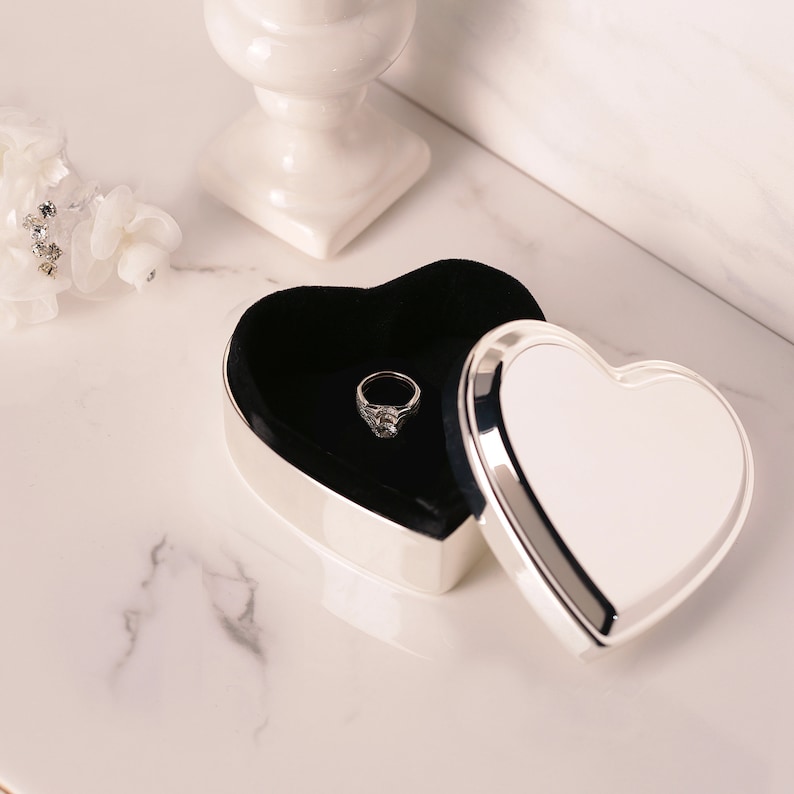 May include: A silver heart-shaped ring box with a black velvet interior. The box is open and a silver ring with a clear stone is inside.