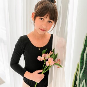 May include: A long-sleeved black leotard featuring a scoop neckline, paired with a delicate gold chain necklace holding a small pendant, and light-colored tights. The garment is worn with a stem of pink alstroemeria flowers held in the hand.