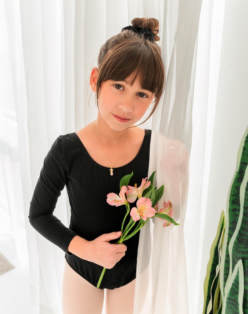 May include: A long-sleeved black leotard featuring a scoop neckline, paired with a delicate gold chain necklace holding a small pendant, and light-colored tights. The garment is worn with a stem of pink alstroemeria flowers held in the hand.