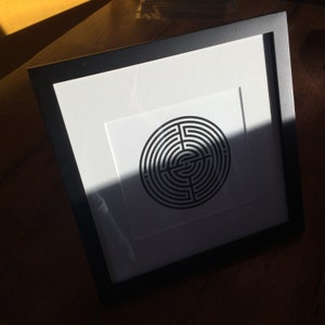 May include: A black and white framed print of a circular maze design. The maze is centered on a white background and has a black frame.
