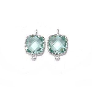 Può includere: Due ciondoli pendenti quadrati color argento con un cristallo verde chiaro al centro. I ciondoli hanno un bordo decorativo e un anello in alto.